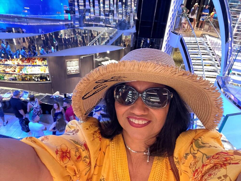 A woman smiling while wearing a sun hat, sunglasses, and a vibrant yellow top while on a cruise ship.